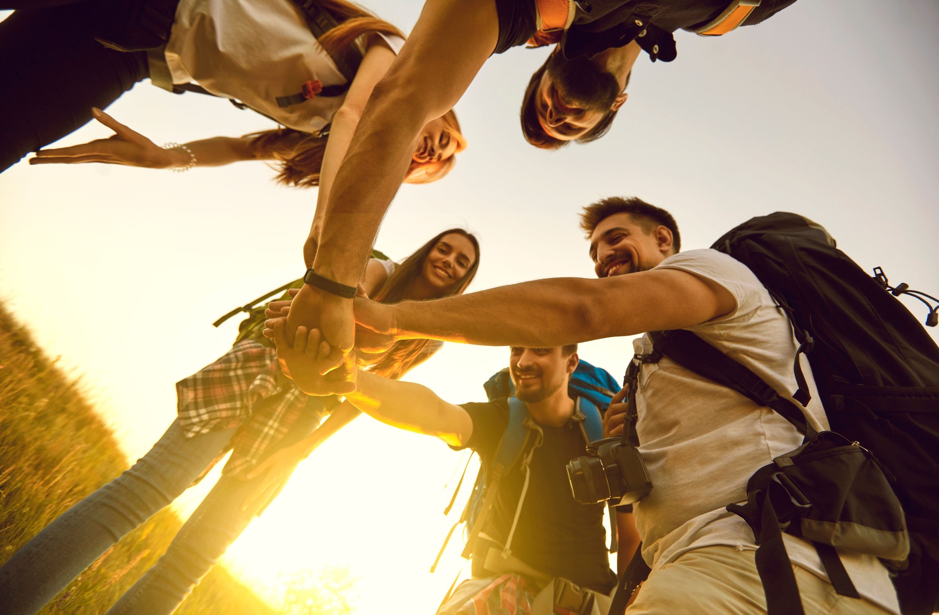 Low angle of team of tourists with rucksacks joining hands in field while hiking in countryside